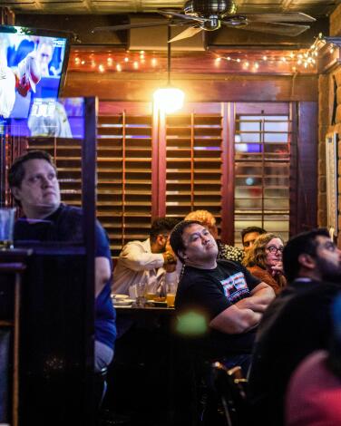 Three people sit at a pub table. One person is clutching his head with both hands.