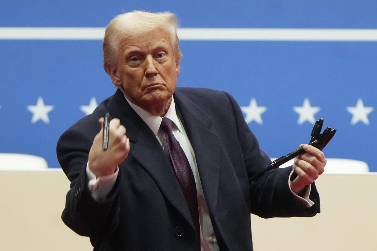A man with blond hair, in a dark suit and tie, throws a black pen to a crowd while holding some in his other hand