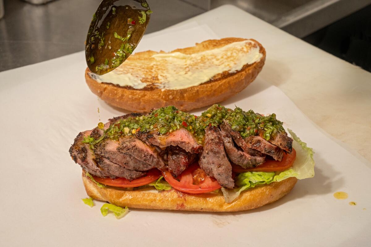 NEWPORT BEACH, CA - OCTOBER 29, 2024: Drizzling chimichurri on the famous steak sandwich at Mario's Butcher Shop in Newport Beach. (Ron De Angelis / For The Times)