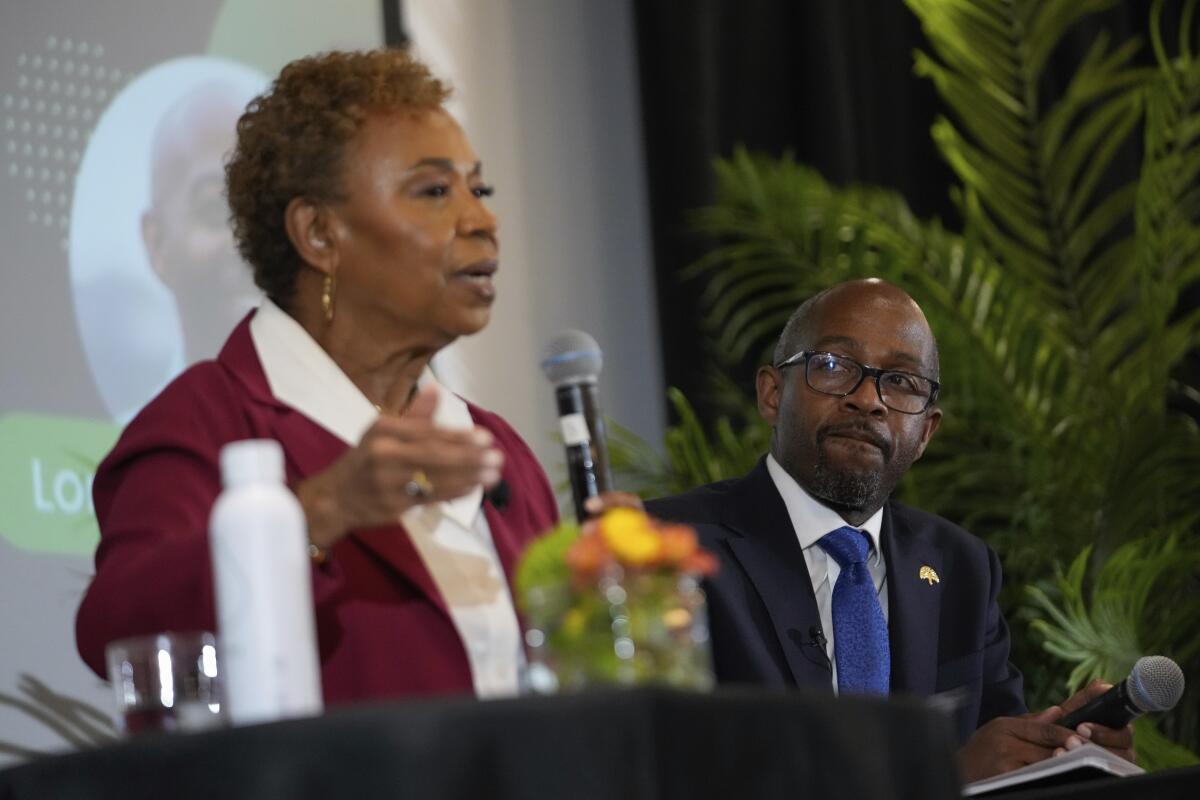 Loren Taylor, in a dark suit, sits next to Barbara Lee, wearing a maroon suit, during a debate in March.