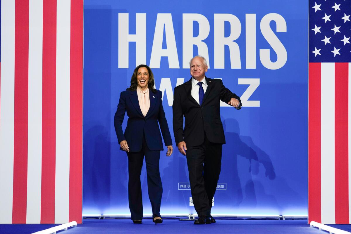Kamala Harris and Tim Walz walk onto a stage before a flag-oriented display that says "Harris Walz"