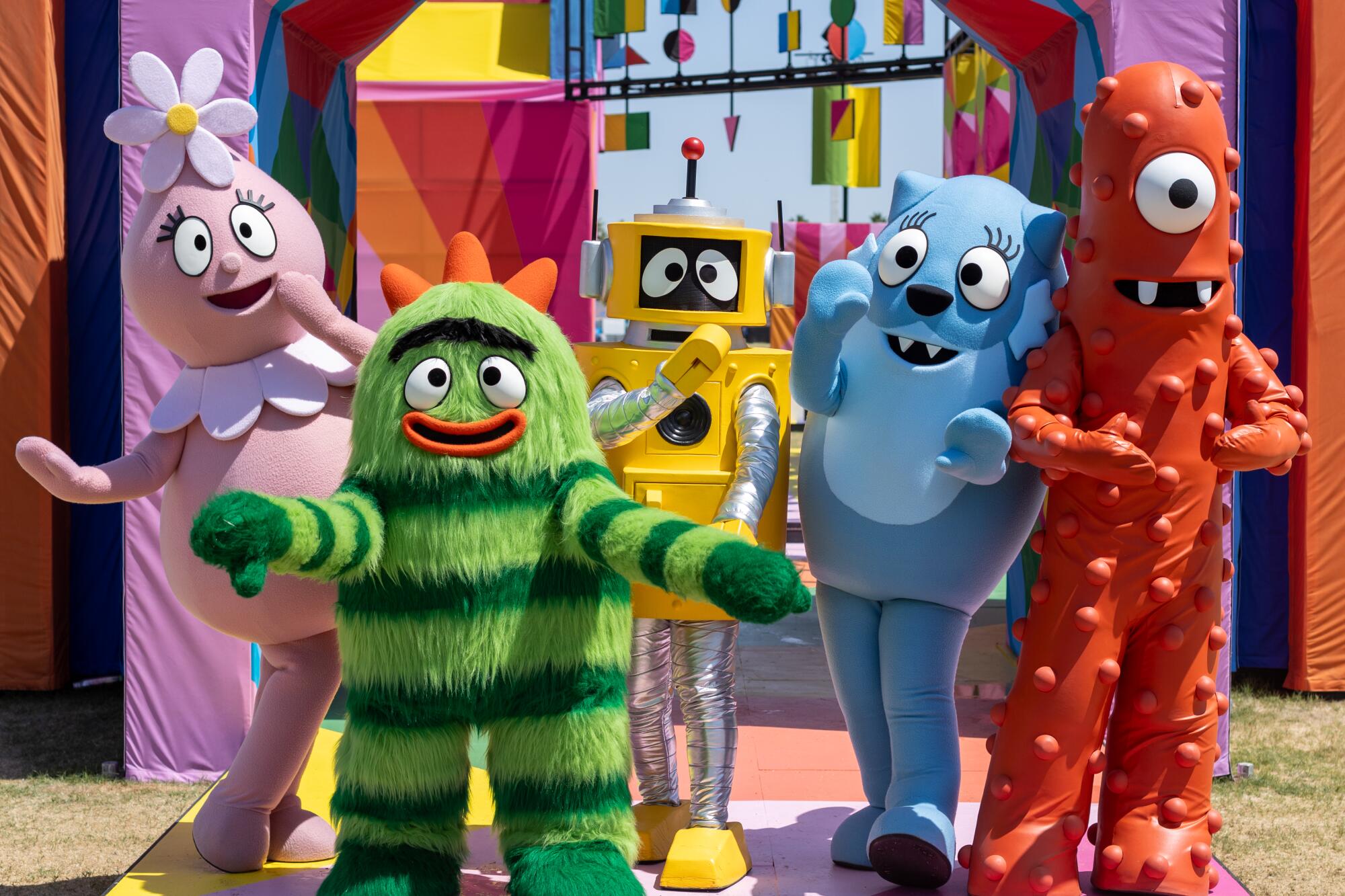 The five brightly colored Yo Gabba Gabba creatures stand in front of a rainbow-colored archway