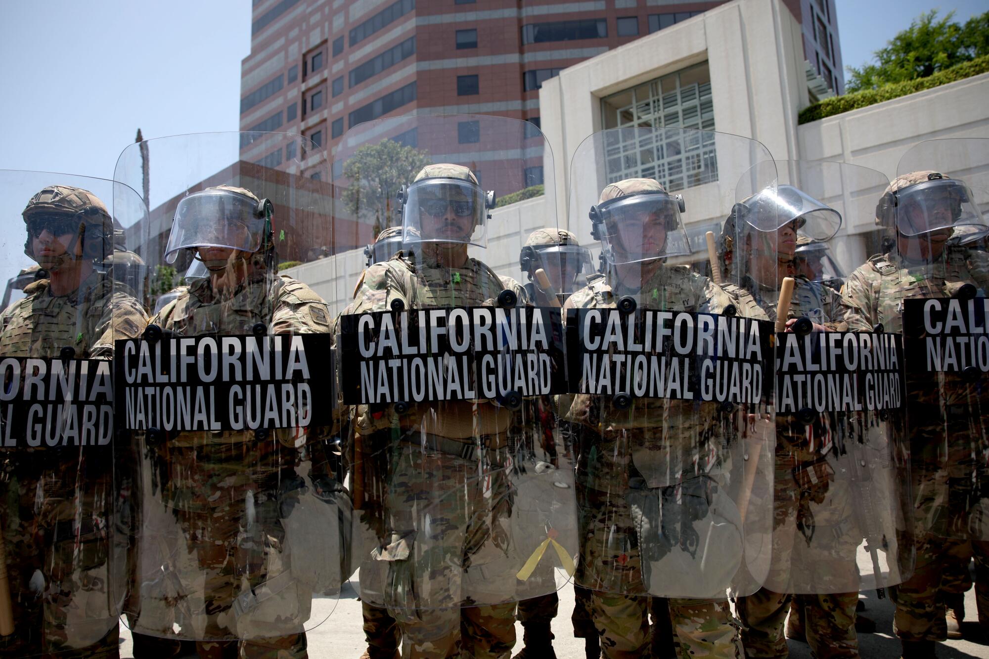 A line of California National Guard tropps in gear with shields stand in front of a tall building.