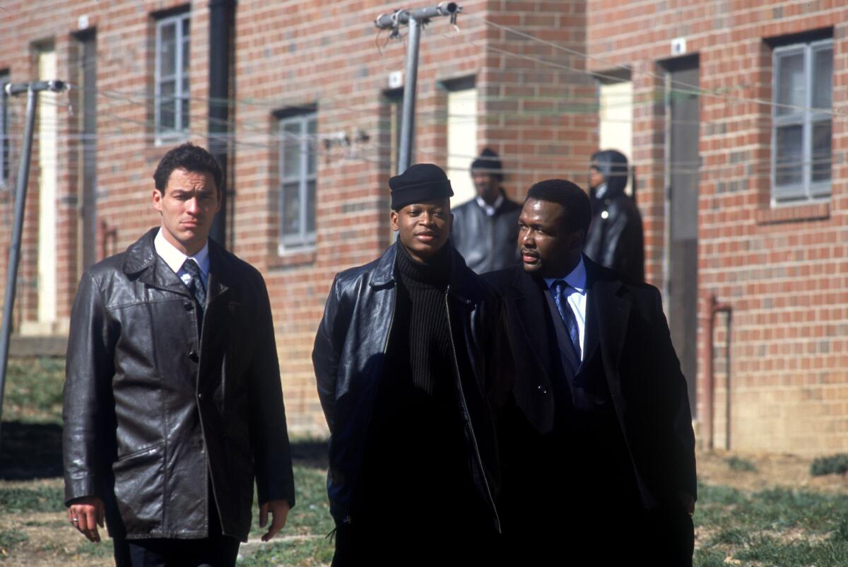 Three men in dark coats walking beside a brick building.
