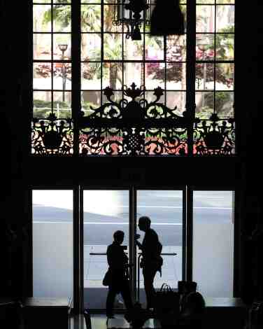 People stand at the glass door of a hotel under ornate windows, with a view out toward the street